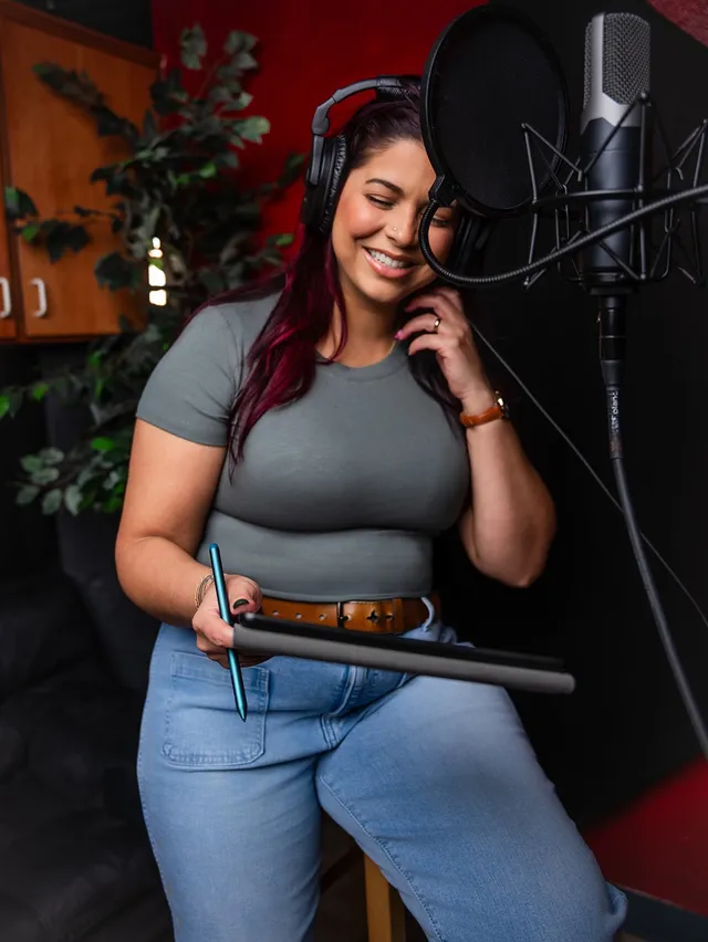 Victoria Rojas recording at a condenser microphone behind a pop filter, headphones on, reading from a tablet