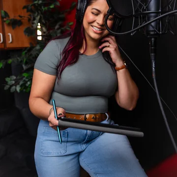 Victoria Rojas recording at a condenser microphone behind a pop filter, headphones on, reading from a tablet