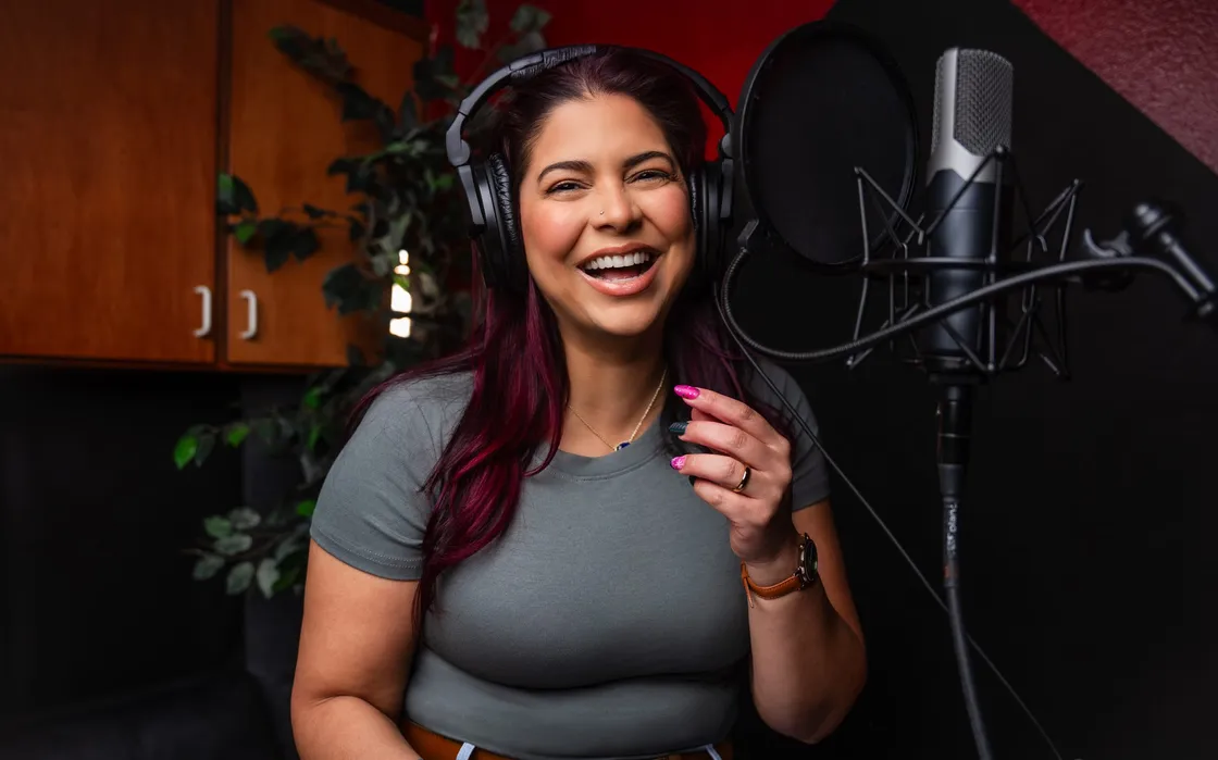Victoria Rojas laughing mid-take at the microphone, headphones on and a script tablet in her hand