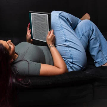 Victoria Rojas stretched out on a couch reading from a backlit e-reader, knees drawn up